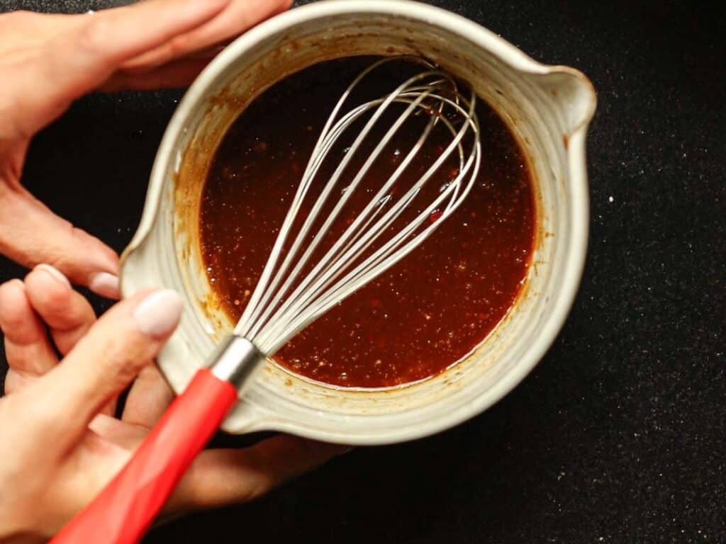 Hands holding a green bowl with reddish-brown sauce being whisked by a red-handled whisk against a black background.